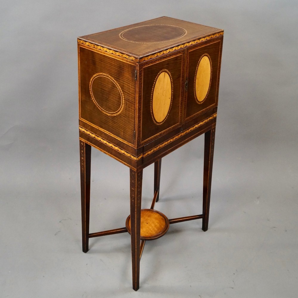 A Sheraton period harewood cabinet on stand featuring twin cupboard doors with oval satinwood inlays and slender tapered legs connected by a circular stretcher tray. View VI