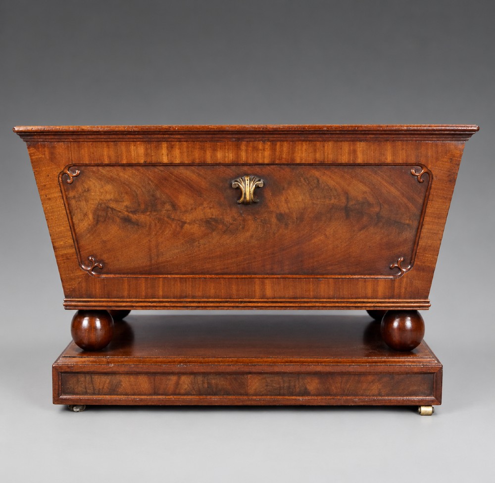 A Regency flame mahogany tapered wine cooler with a central brass escutcheon, supported by turned spherical feet and a rectangular plinth base.