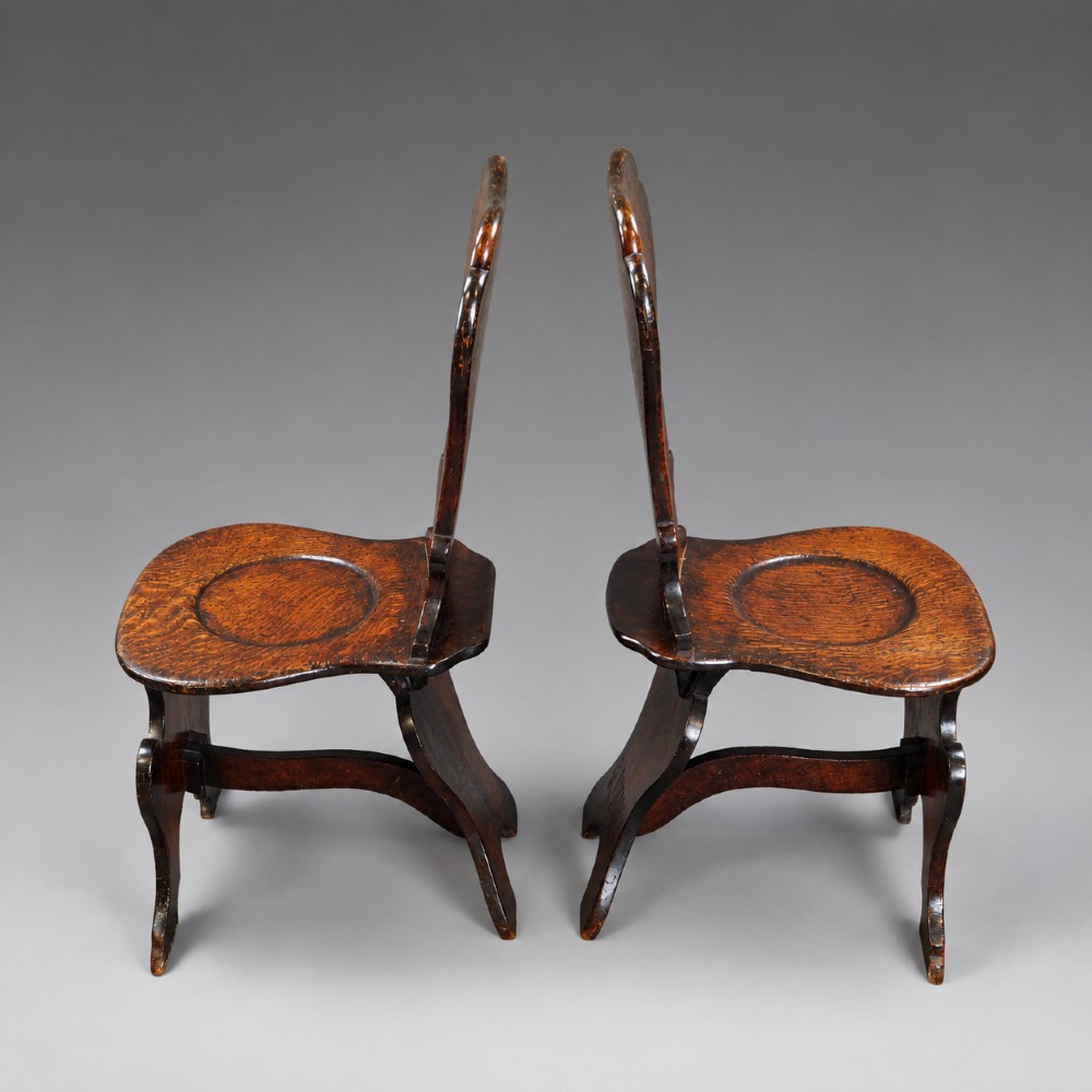 A pair of dark oak sgabello hall chairs featuring contoured seats, scroll-cut trestle bases, and shaped cartouche backrests with painted green and gold central roundels. View II
