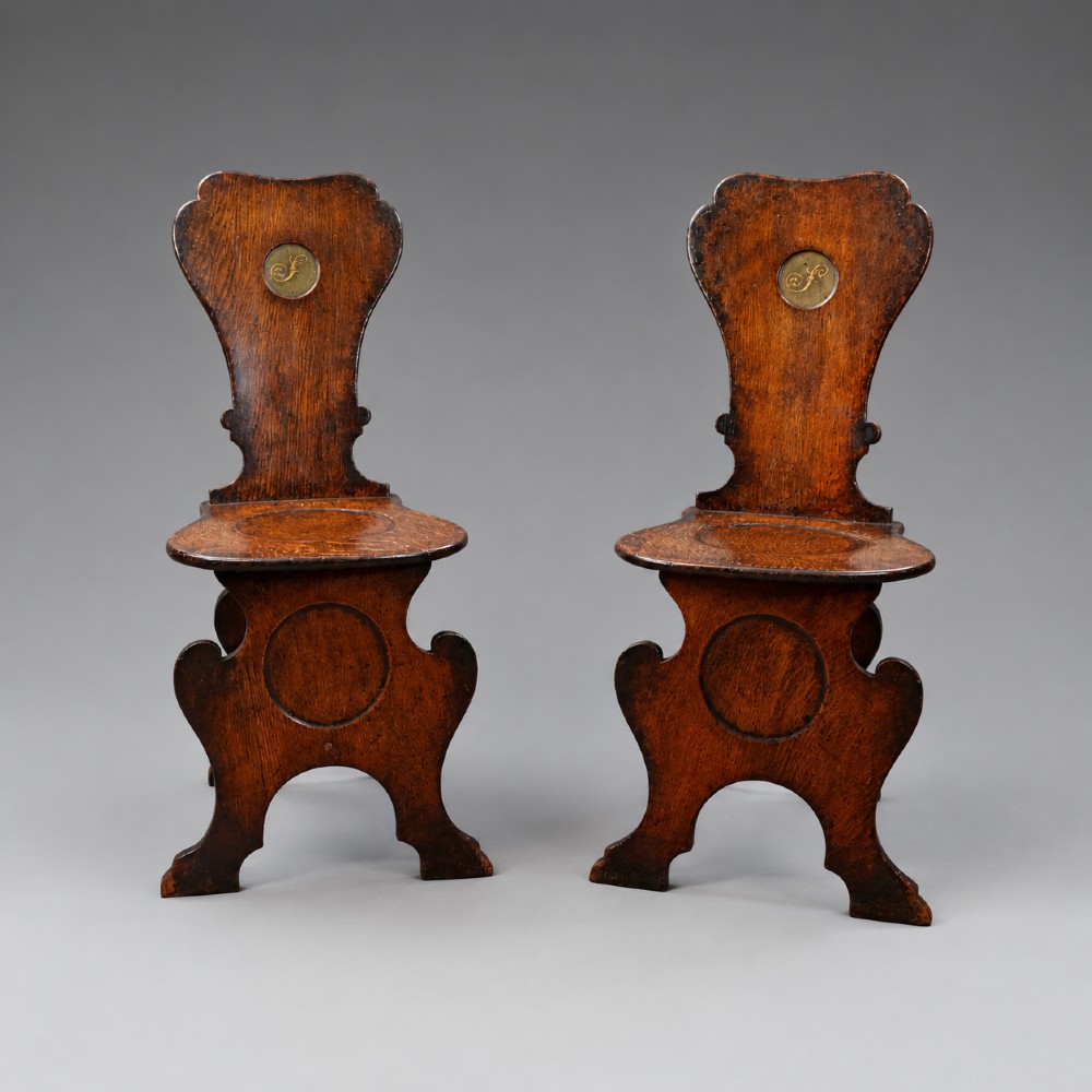 A pair of dark oak sgabello hall chairs featuring contoured seats, scroll-cut trestle bases, and shaped cartouche backrests with painted green and gold central roundels. View IV