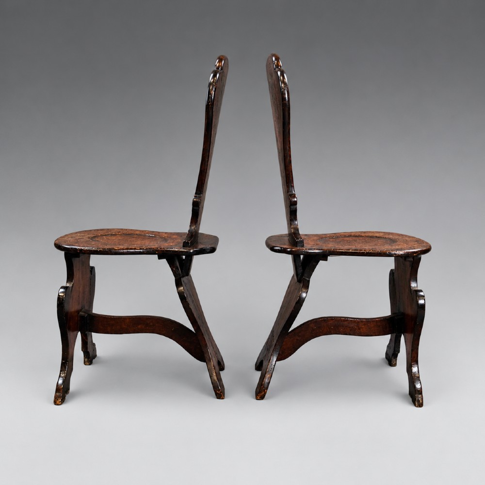 A pair of dark oak sgabello hall chairs featuring contoured seats, scroll-cut trestle bases, and shaped cartouche backrests with painted green and gold central roundels. View V