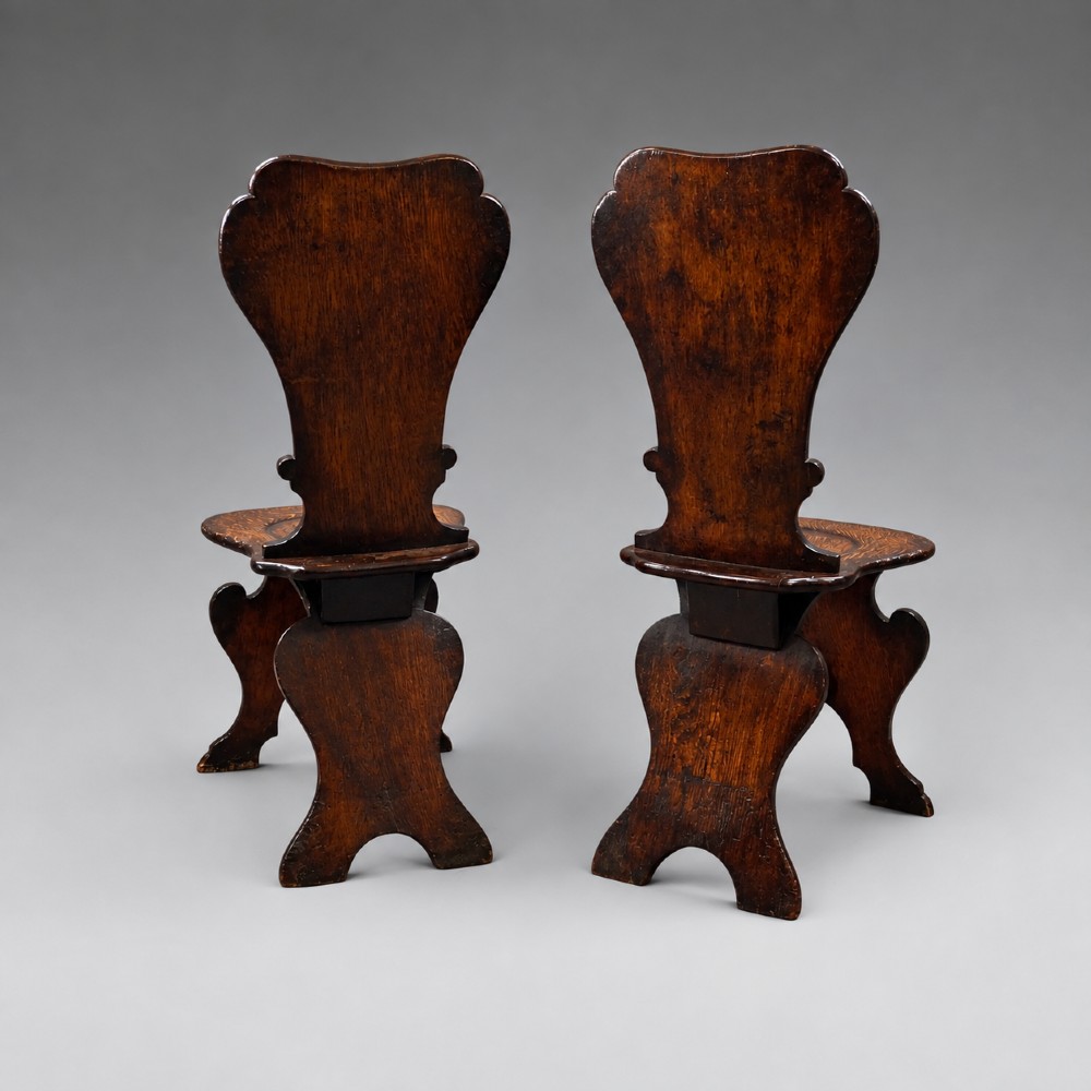 A pair of dark oak sgabello hall chairs featuring contoured seats, scroll-cut trestle bases, and shaped cartouche backrests with painted green and gold central roundels. View VI