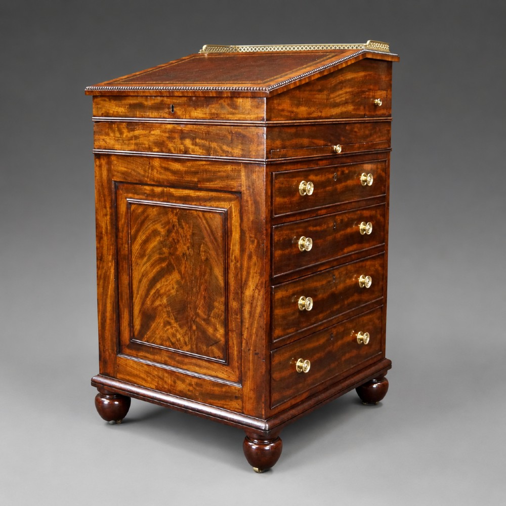 An antique flame mahogany Davenport desk featuring a slanted leather writing surface, perforated brass gallery, side drawers with turned brass knob handles, and compressed bun feet.