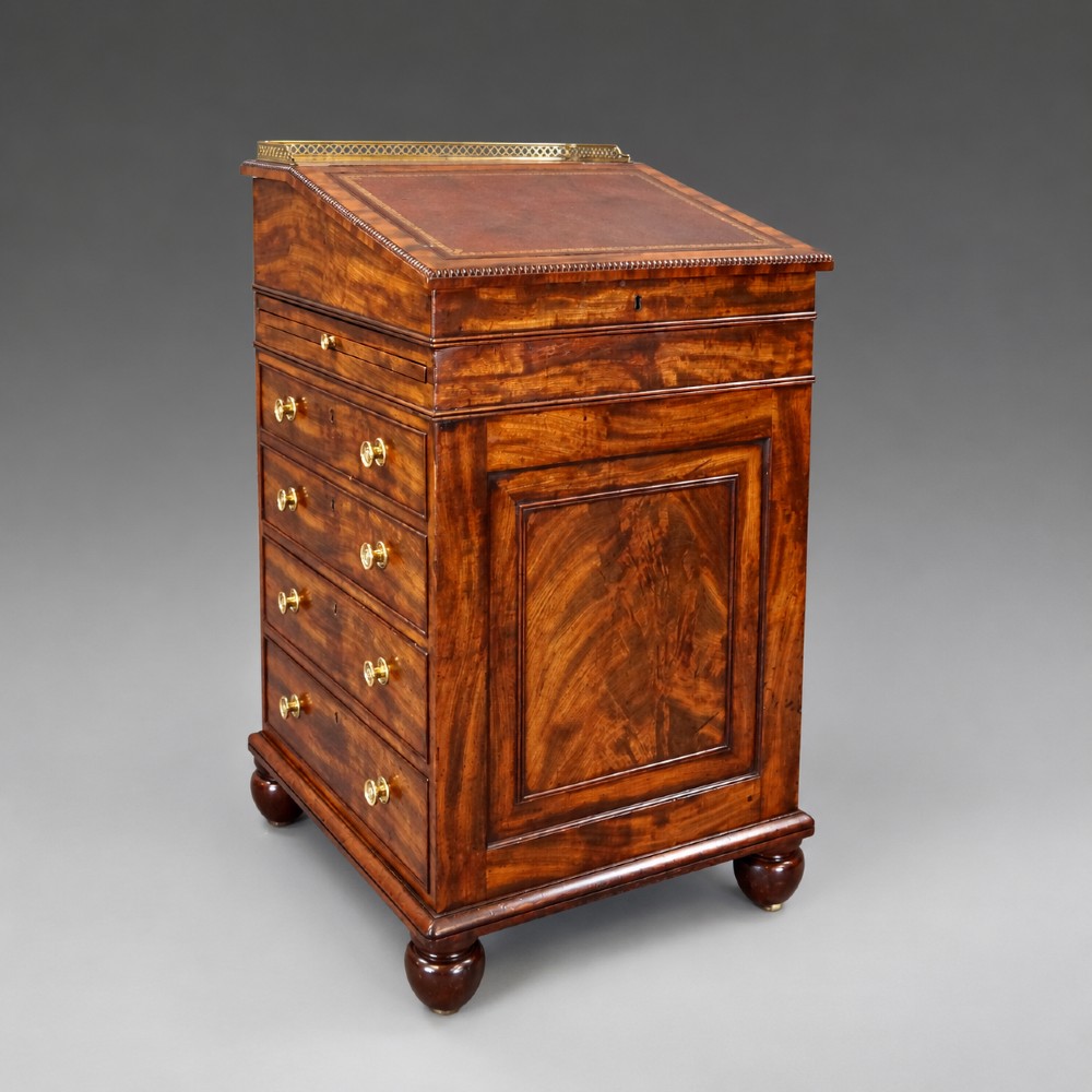 An antique flame mahogany Davenport desk featuring a slanted leather writing surface, perforated brass gallery, side drawers with turned brass knob handles, and compressed bun feet. View II