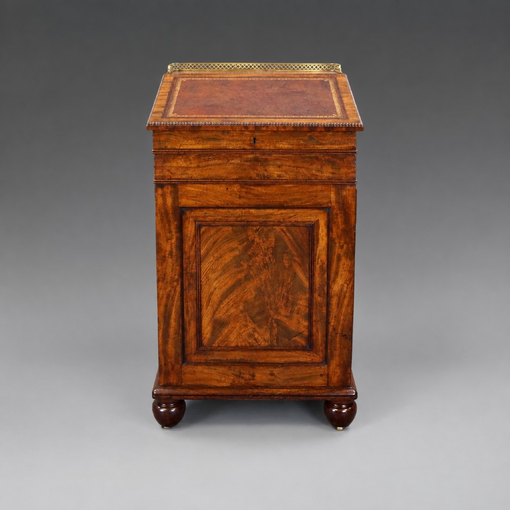 An antique flame mahogany Davenport desk featuring a slanted leather writing surface, perforated brass gallery, side drawers with turned brass knob handles, and compressed bun feet. View III