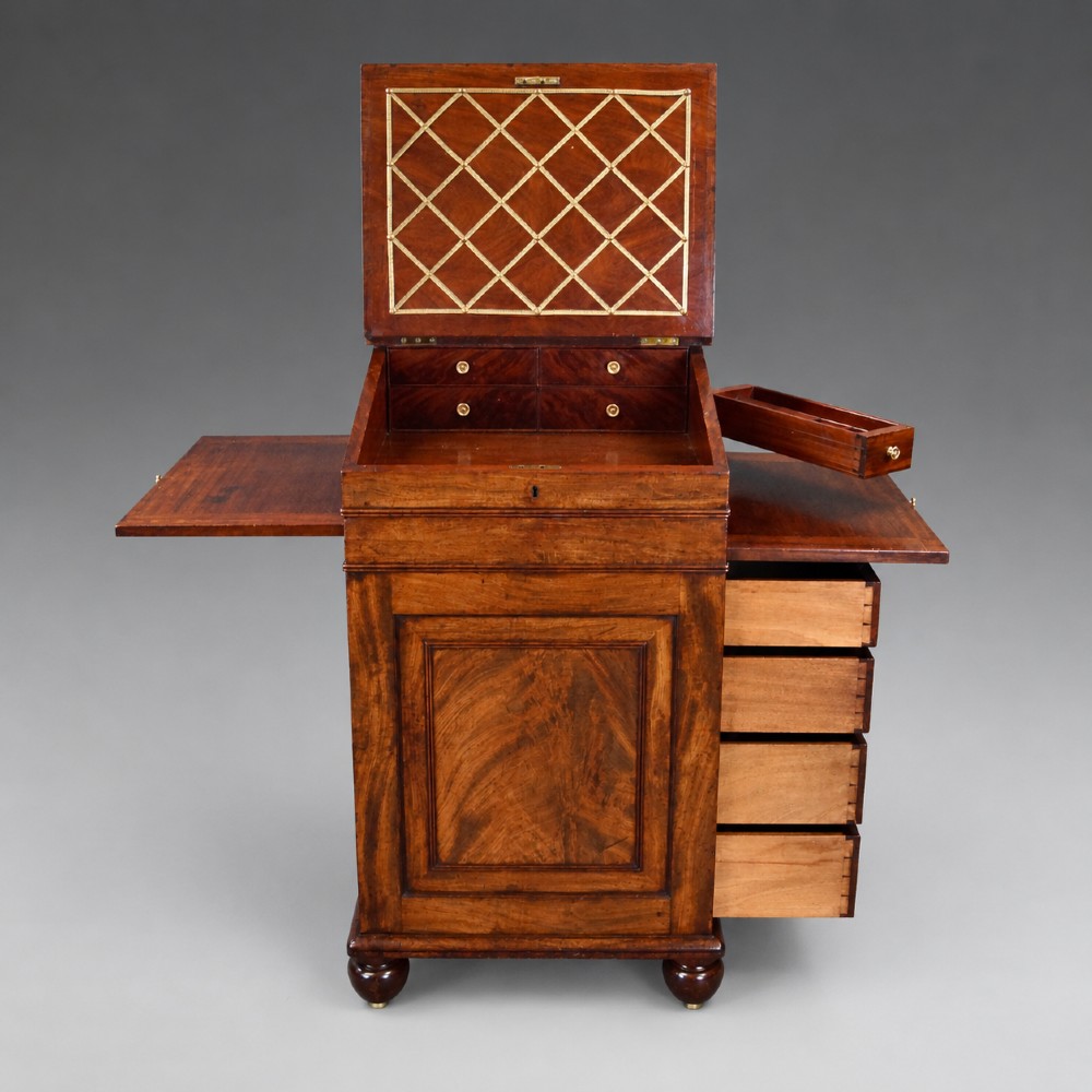 An antique flame mahogany Davenport desk featuring a slanted leather writing surface, perforated brass gallery, side drawers with turned brass knob handles, and compressed bun feet. View IV