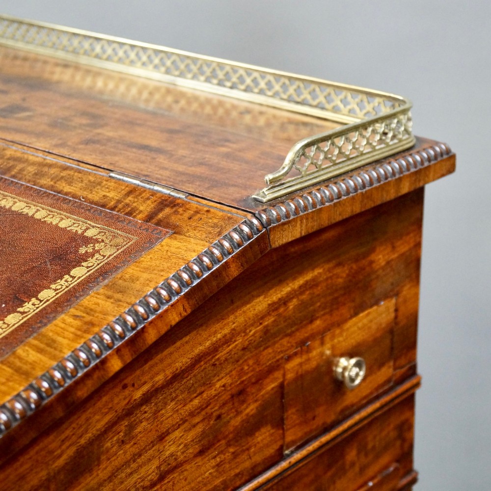 An antique flame mahogany Davenport desk featuring a slanted leather writing surface, perforated brass gallery, side drawers with turned brass knob handles, and compressed bun feet. View V