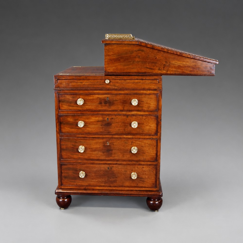 An antique flame mahogany Davenport desk featuring a slanted leather writing surface, perforated brass gallery, side drawers with turned brass knob handles, and compressed bun feet. View VI