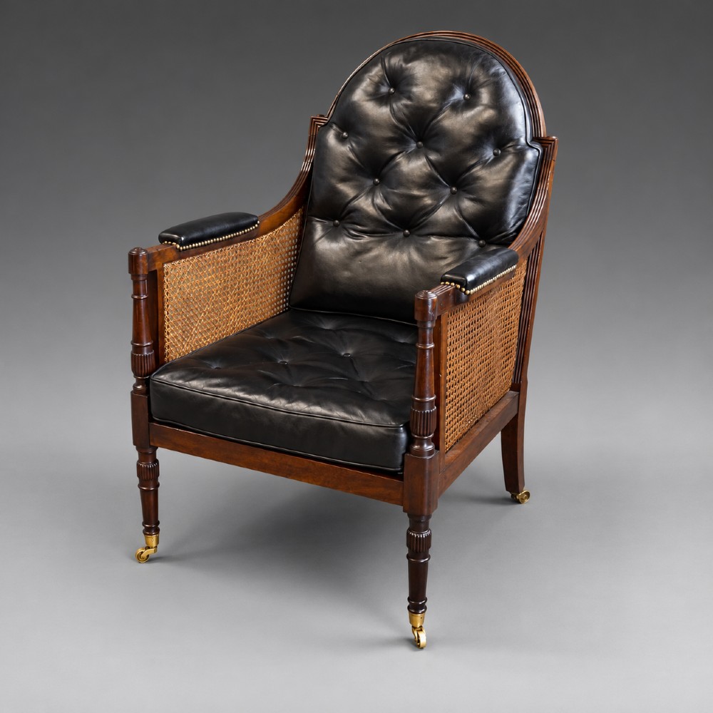 A Regency mahogany bergere armchair featuring woven cane side panels, button-tufted black leather upholstery, reeded turned front legs, and brass castors.