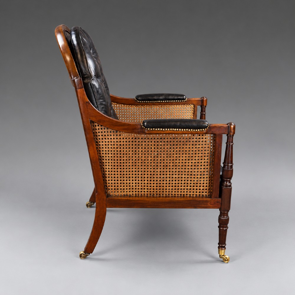 A Regency mahogany bergere armchair featuring woven cane side panels, button-tufted black leather upholstery, reeded turned front legs, and brass castors. View III