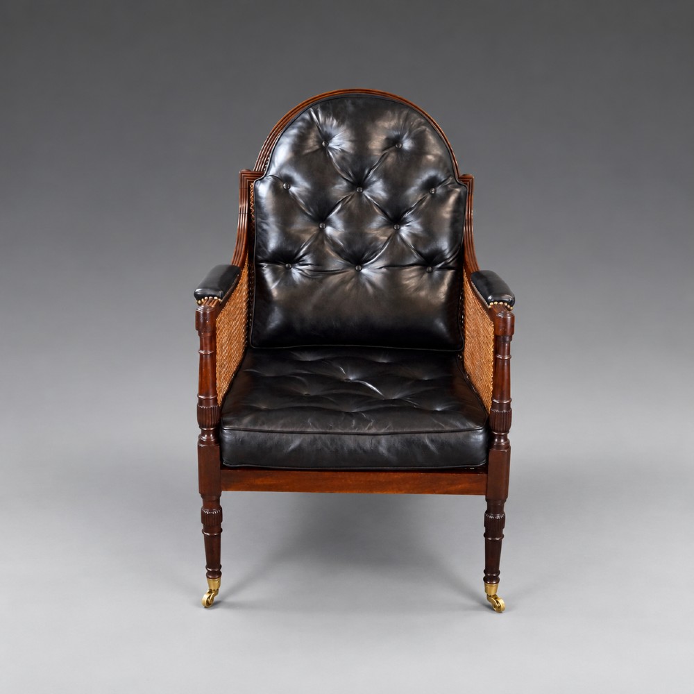 A Regency mahogany bergere armchair featuring woven cane side panels, button-tufted black leather upholstery, reeded turned front legs, and brass castors. View IV