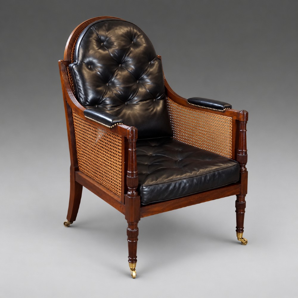 A Regency mahogany bergere armchair featuring woven cane side panels, button-tufted black leather upholstery, reeded turned front legs, and brass castors. View V