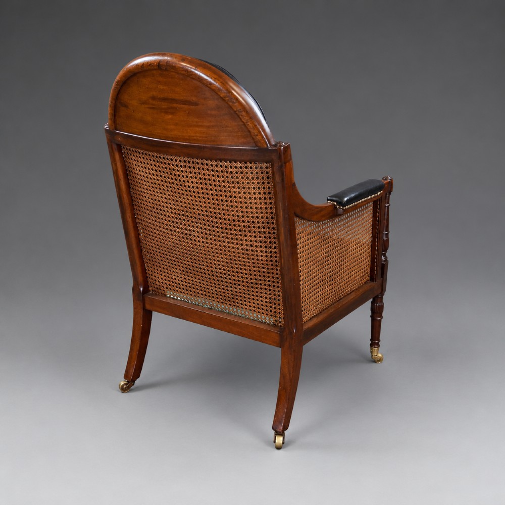 A Regency mahogany bergere armchair featuring woven cane side panels, button-tufted black leather upholstery, reeded turned front legs, and brass castors. View VI