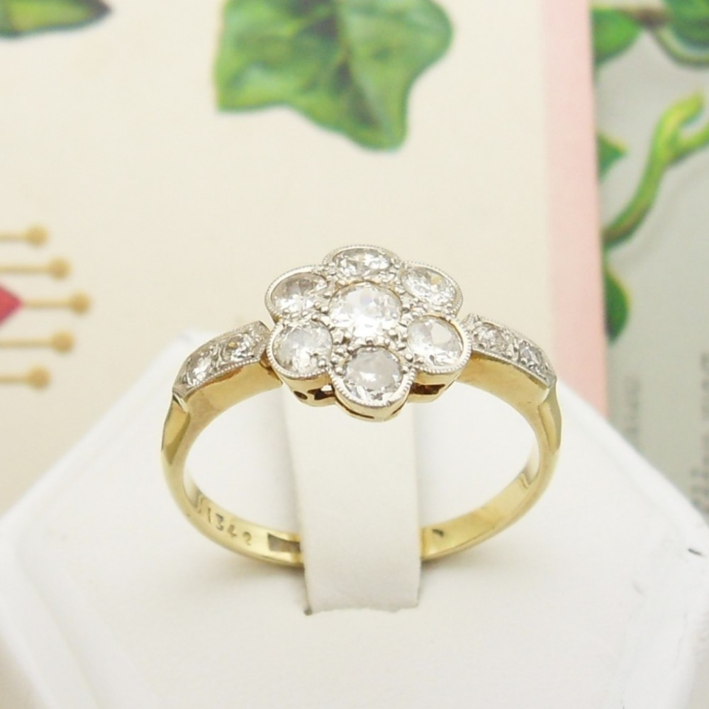 An Edwardian 18ct gold and platinum ring featuring a flower-shaped diamond cluster setting with millegrain edges and diamond-set shoulders.