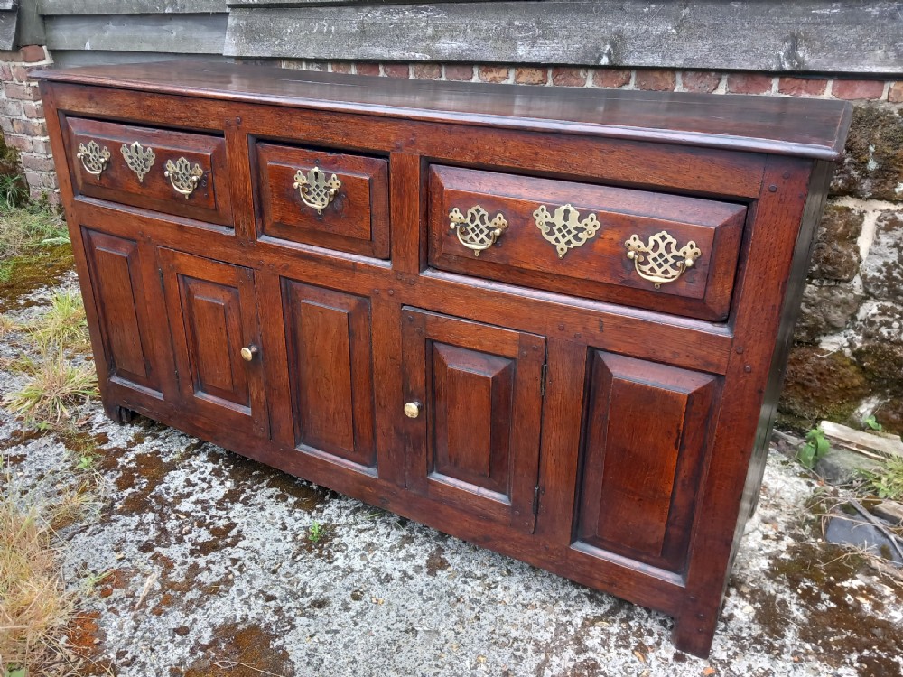 An eighteenth-century dark oak dresser base featuring three drawers with pierced brass handles situated above panelled cupboard doors. View II