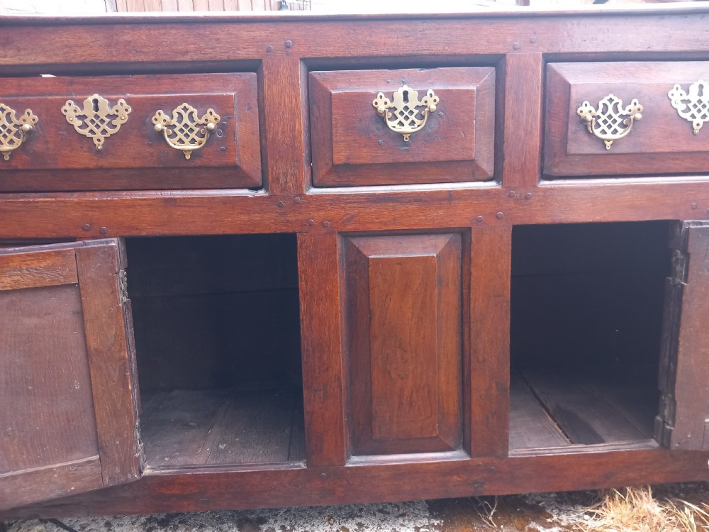 An eighteenth-century dark oak dresser base featuring three drawers with pierced brass handles situated above panelled cupboard doors. View V
