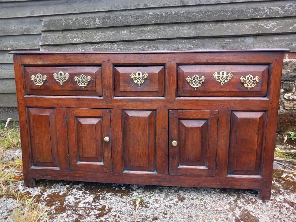 An eighteenth-century dark oak dresser base featuring three drawers with pierced brass handles situated above panelled cupboard doors. View IX