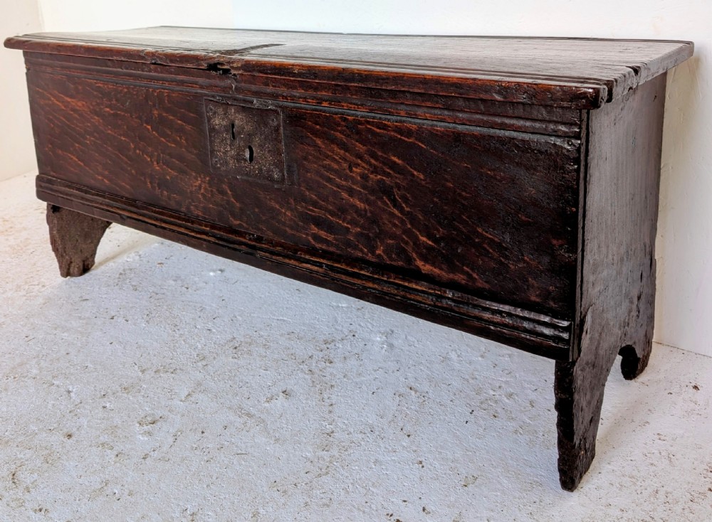 A small 17th-century dark oak plank coffer featuring a hinged rectangular lid, iron escutcheon plate, and V-shaped end supports.