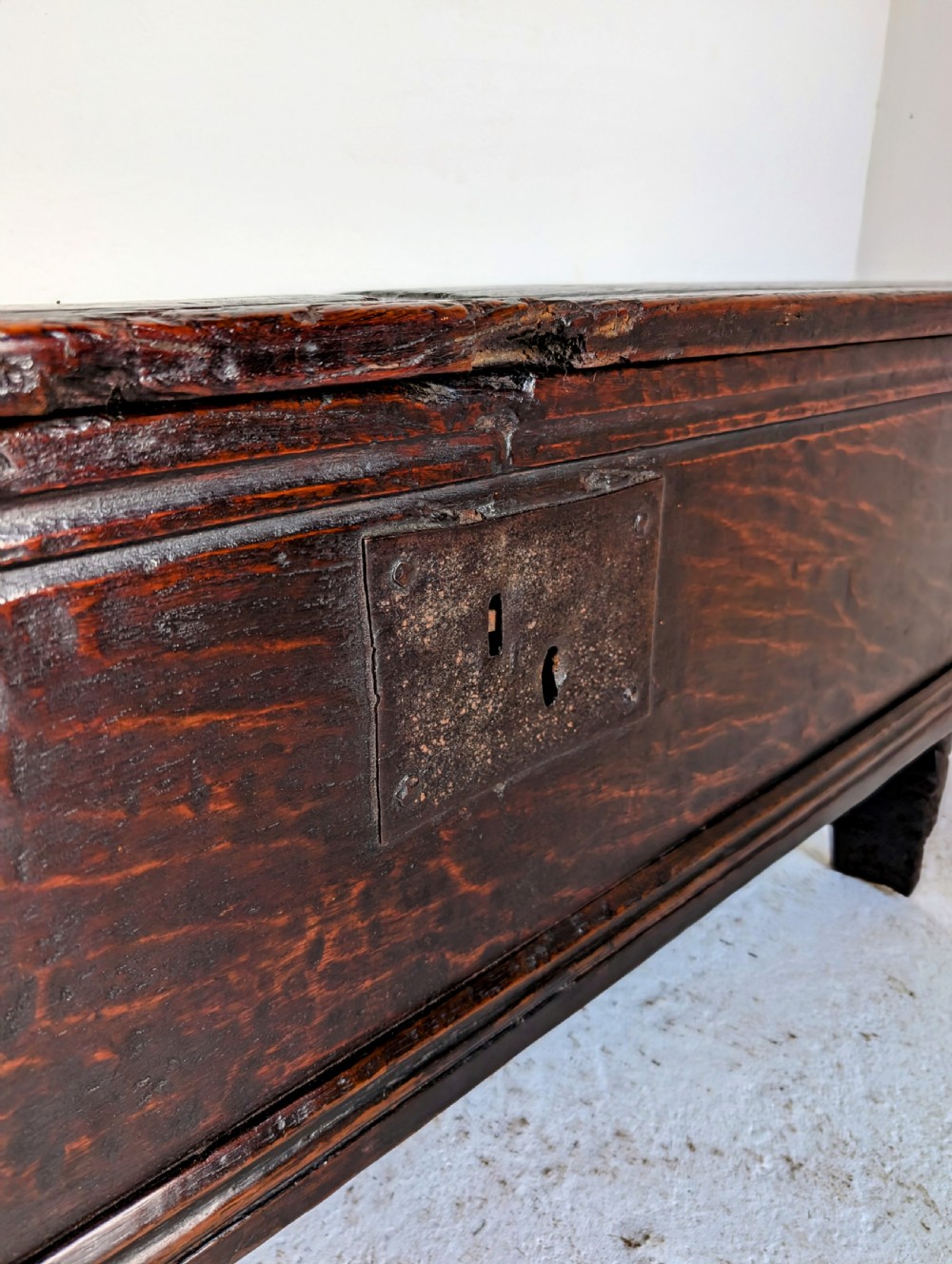 A small 17th-century dark oak plank coffer featuring a hinged rectangular lid, iron escutcheon plate, and V-shaped end supports. View III