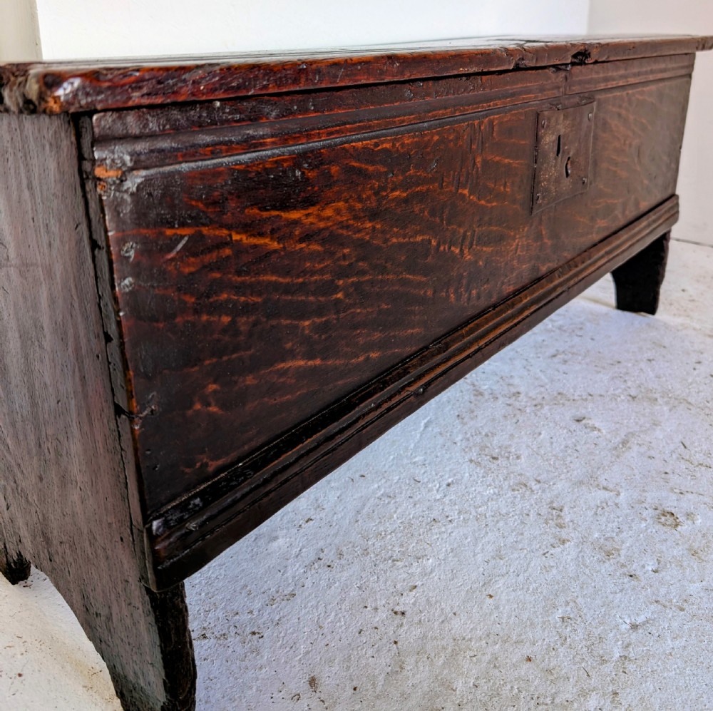 A small 17th-century dark oak plank coffer featuring a hinged rectangular lid, iron escutcheon plate, and V-shaped end supports. View V