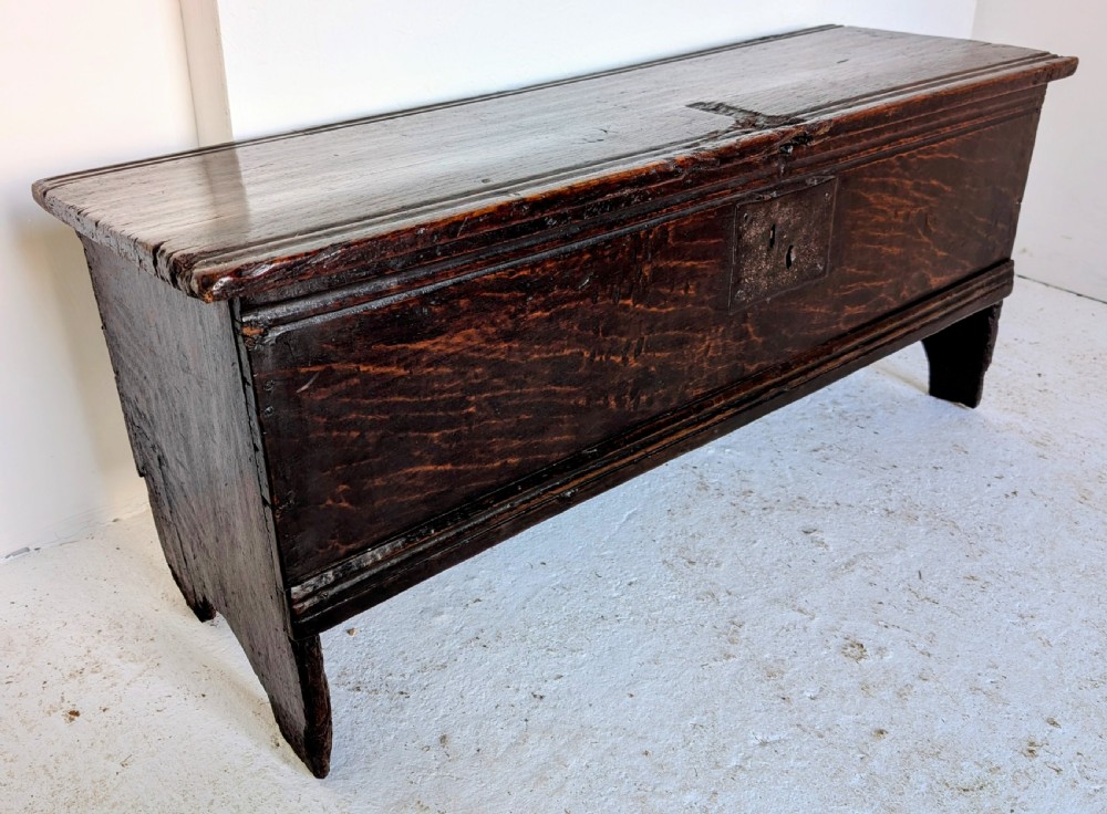 A small 17th-century dark oak plank coffer featuring a hinged rectangular lid, iron escutcheon plate, and V-shaped end supports. View VI