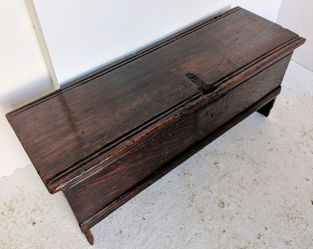A small 17th-century dark oak plank coffer featuring a hinged rectangular lid, iron escutcheon plate, and V-shaped end supports. View VII
