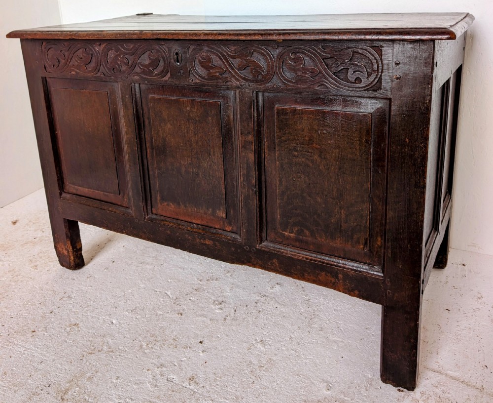 A dark oak 17th-century coffer featuring three front panels and a carved scrollwork frieze below the hinged lid.