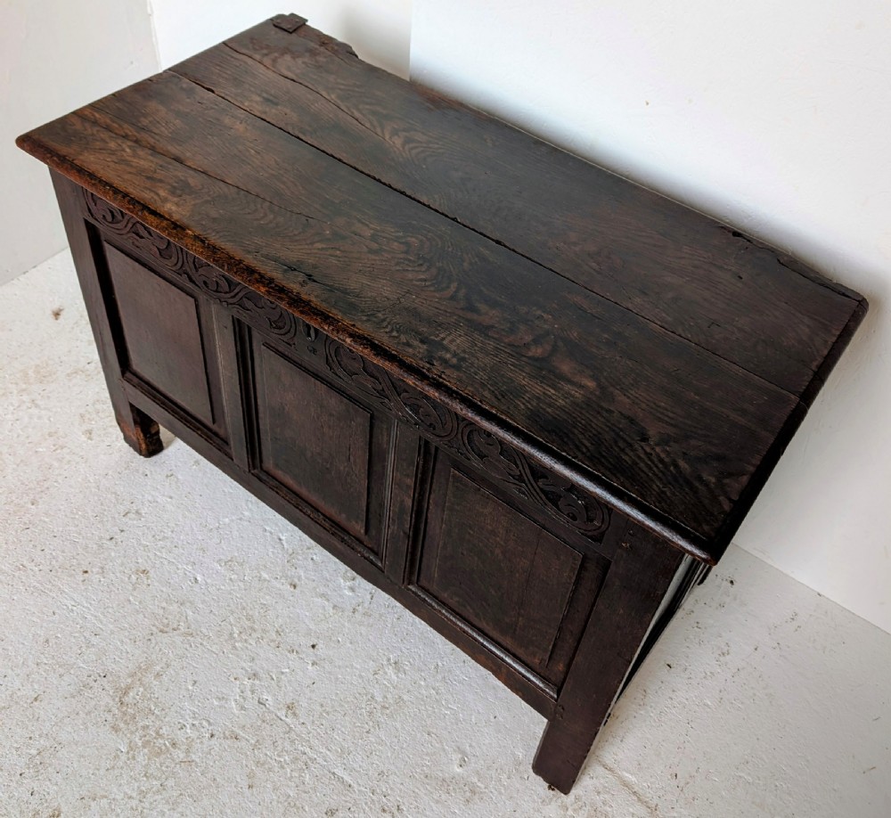 A dark oak 17th-century coffer featuring three front panels and a carved scrollwork frieze below the hinged lid. View II