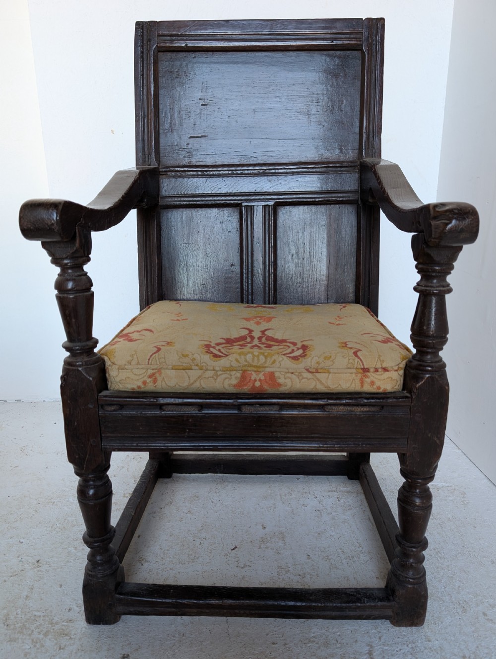 A dark oak 17th-century wainscot armchair featuring a panelled backrest, turned front legs, robust armrests, and an upholstered seat cushion.