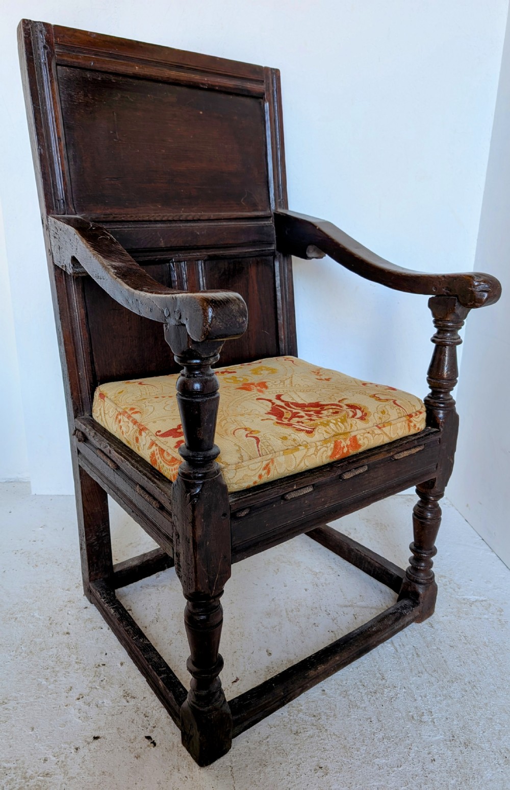 A dark oak 17th-century wainscot armchair featuring a panelled backrest, turned front legs, robust armrests, and an upholstered seat cushion. View II