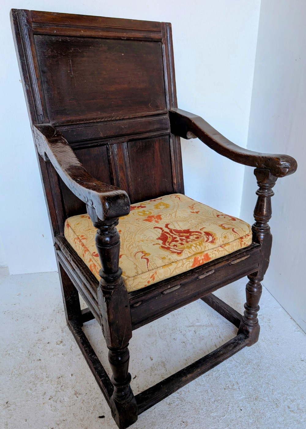 A dark oak 17th-century wainscot armchair featuring a panelled backrest, turned front legs, robust armrests, and an upholstered seat cushion. View IV