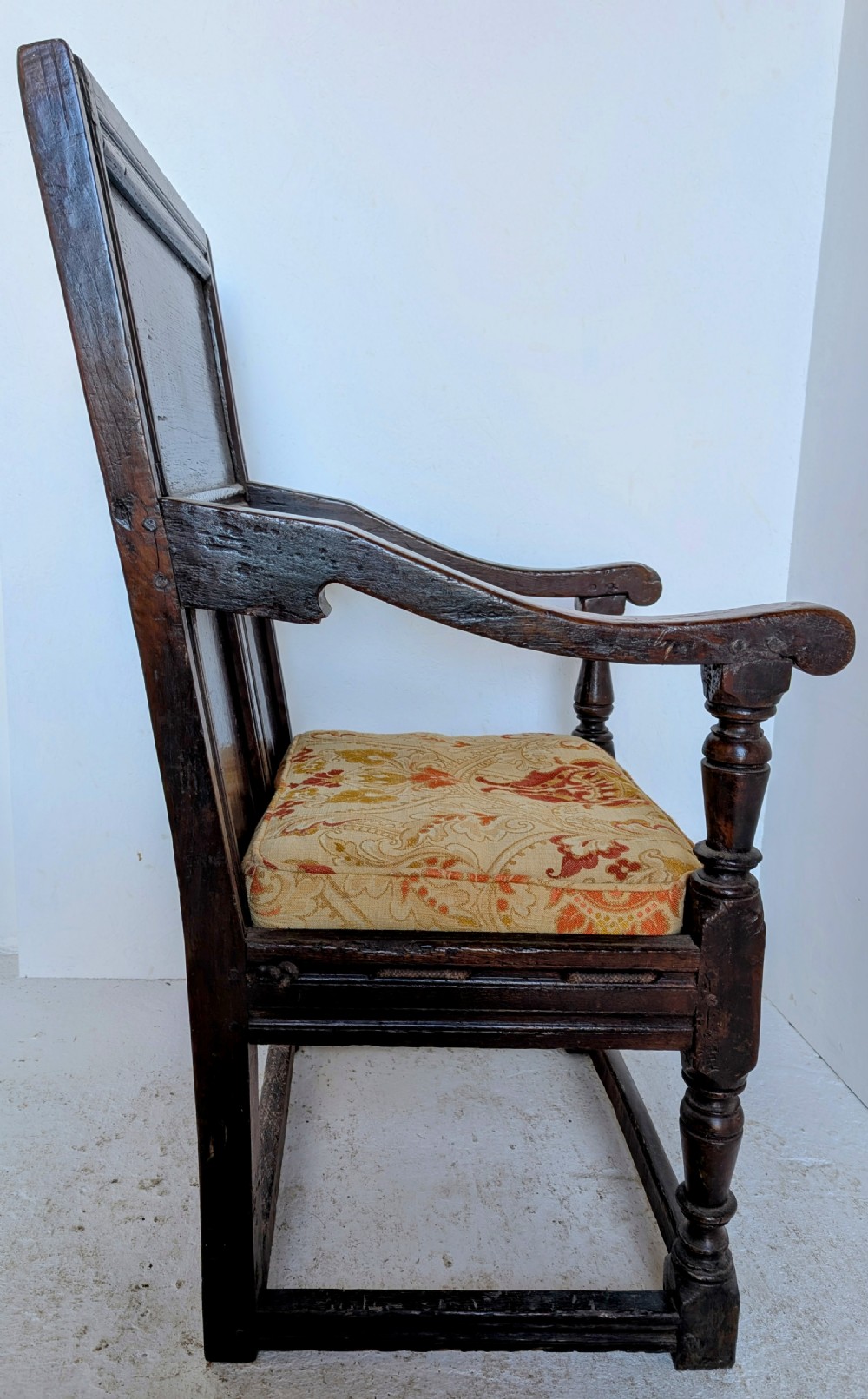 A dark oak 17th-century wainscot armchair featuring a panelled backrest, turned front legs, robust armrests, and an upholstered seat cushion. View VII