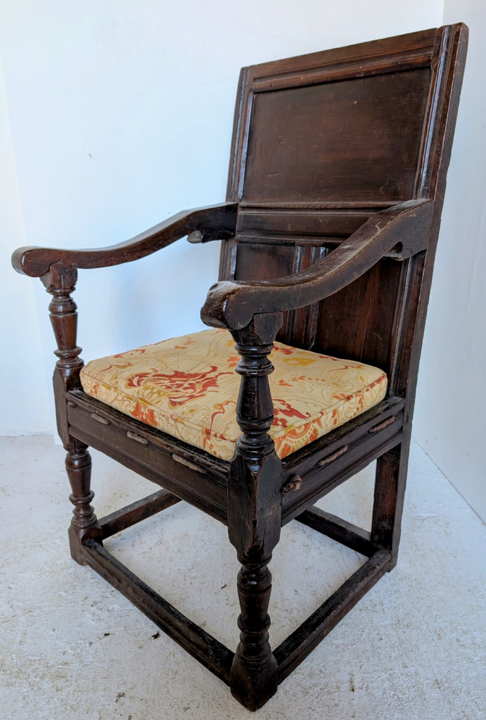A dark oak 17th-century wainscot armchair featuring a panelled backrest, turned front legs, robust armrests, and an upholstered seat cushion. View IX