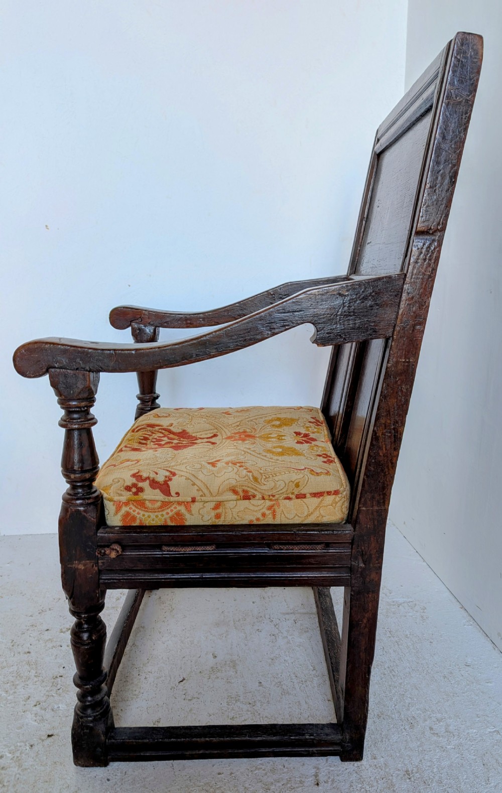 A dark oak 17th-century wainscot armchair featuring a panelled backrest, turned front legs, robust armrests, and an upholstered seat cushion. View X