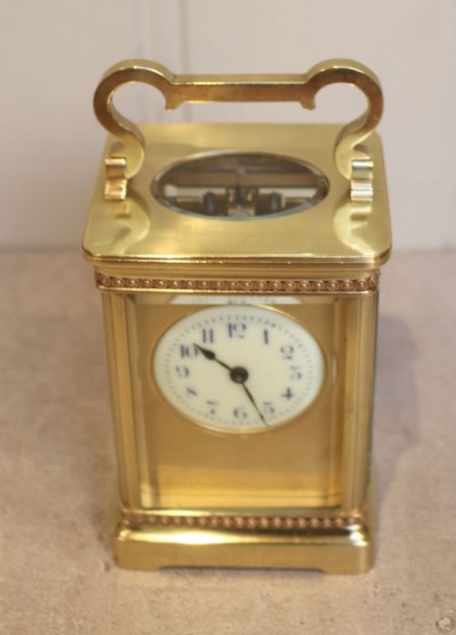 fig 1. An Edwardian brass carriage clock featuring a rectangular case with glass panels, beaded trim, a top handle, and a white enamel dial with blue numerals.