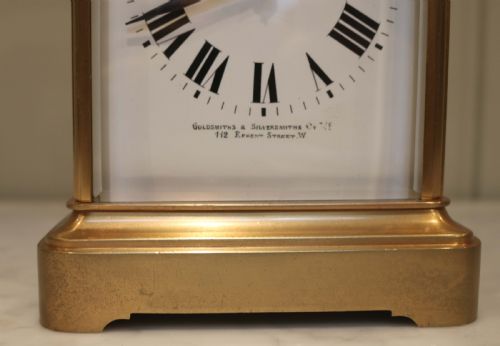fig 1. A large rectangular brass carriage clock featuring bevelled glass side panels, a white enamel dial with black Roman numerals, and a hinged top handle.