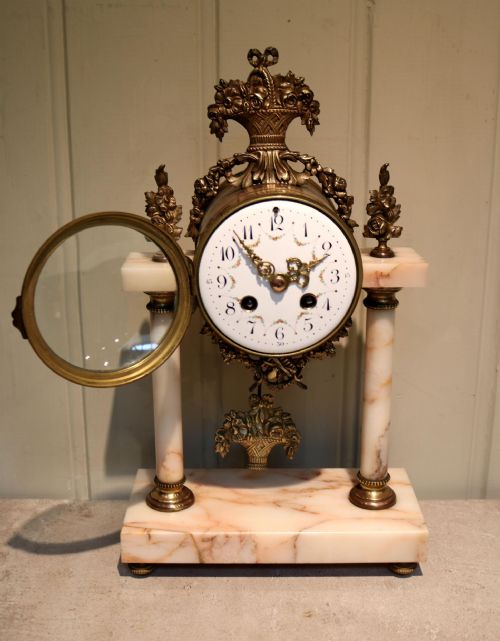 fig 1. A three-piece French clock garniture set constructed from pale pink marble and gilt brass, featuring a central portico clock with a white enamel dial flanked by two matching decorative urns.