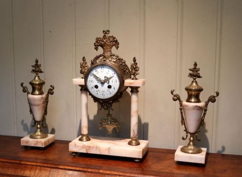 fig 2. A three-piece French clock garniture set constructed from pale pink marble and gilt brass, featuring a central portico clock with a white enamel dial flanked by two matching decorative urns.