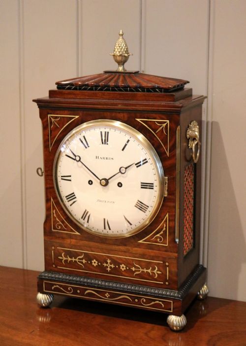 fig 1. A Regency flame mahogany bracket clock with detailed brass inlay, a white circular dial signed Harris Brixton, side ring handles, and a brass pineapple finial.