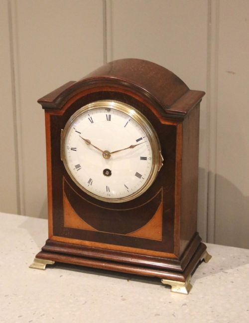 fig 1. An arched mahogany mantel clock featuring geometric satinwood inlay, a white dial with Roman numerals, a brass bezel, and small brass feet.