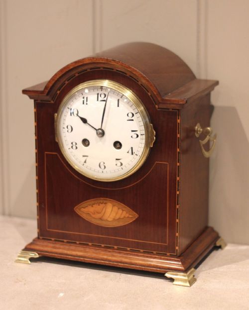 fig 1. A mahogany break-arch mantel clock featuring a shell marquetry inlay, brass bracket feet, side drop handles, and an enamel dial with Arabic numerals.