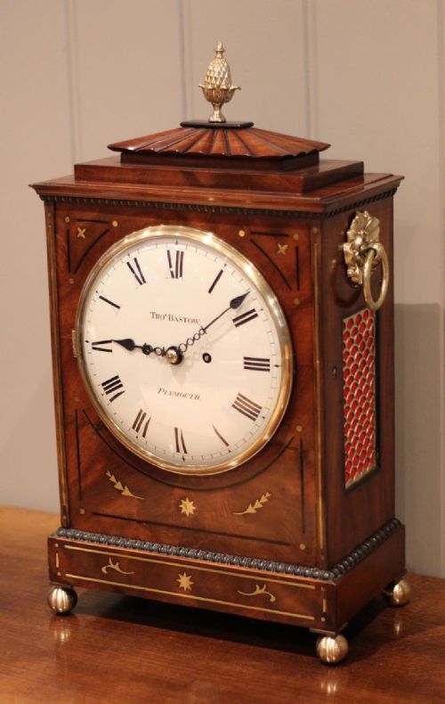 fig 1. A Regency flame mahogany bracket clock featuring brass stringing inlay, side ring handles, a brass finial, and a white circular dial with Roman numerals.
