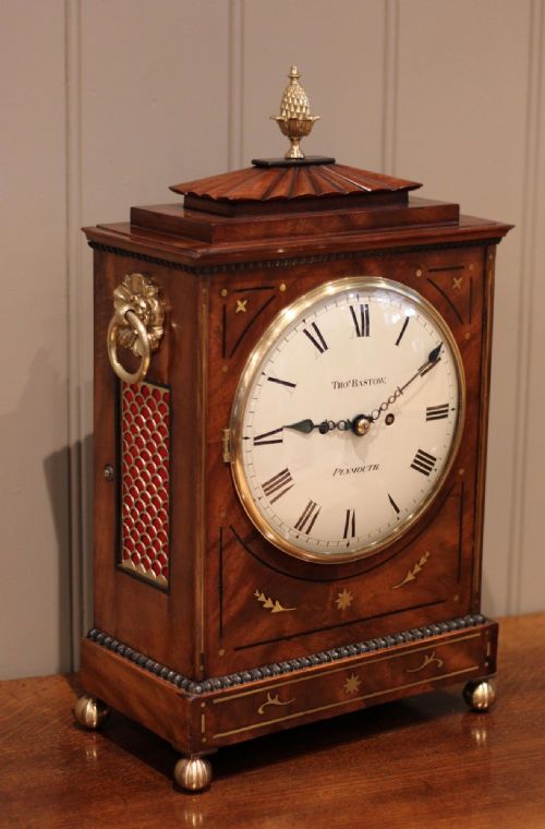 fig 2. A Regency flame mahogany bracket clock featuring brass stringing inlay, side ring handles, a brass finial, and a white circular dial with Roman numerals.