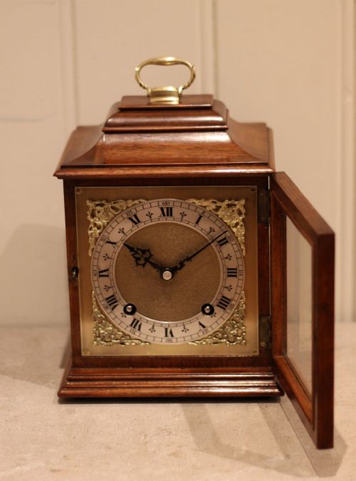 fig 1. A walnut bracket clock featuring a brass carrying handle on top, a clear glass door, and a silvered dial with black Roman numerals and ornate brass spandrels.