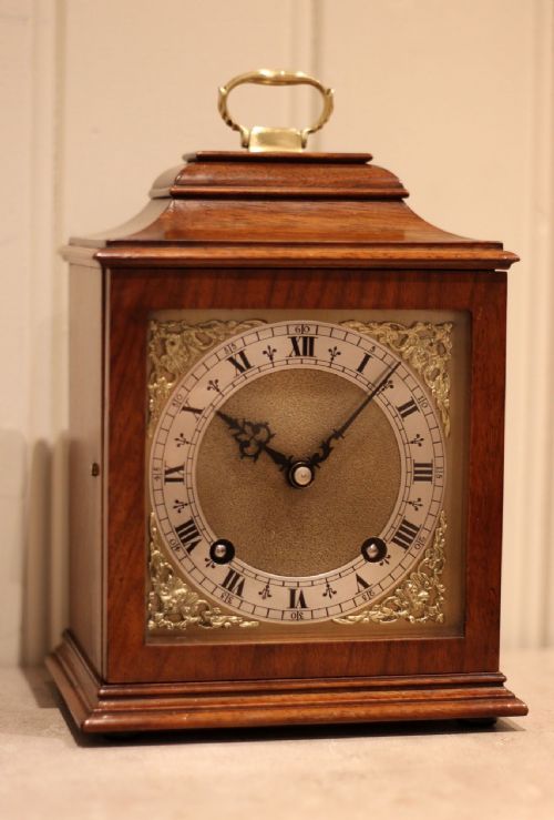 fig 2. A walnut bracket clock featuring a brass carrying handle on top, a clear glass door, and a silvered dial with black Roman numerals and ornate brass spandrels.