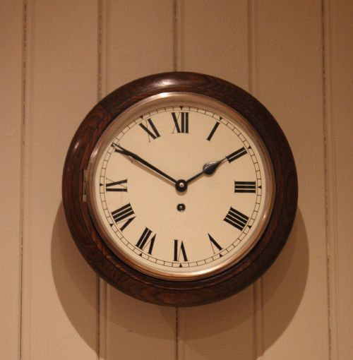 fig 1. A circular early 20th-century wall clock featuring a moulded dark oak frame, a brass bezel, and a white dial face with black Roman numerals.
