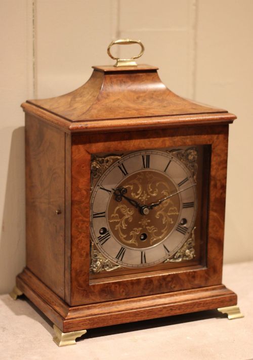 fig 1. A burr walnut bracket clock featuring a caddy top with a brass carry handle, an ornate brass dial with a silvered chapter ring, and brass bracket feet.