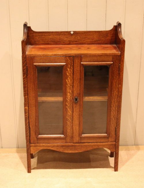 fig 1. A small Arts and Crafts medium oak glazed bookcase featuring a gallery top with a pierced heart motif, two glazed doors, glazed side panels, and square legs.
