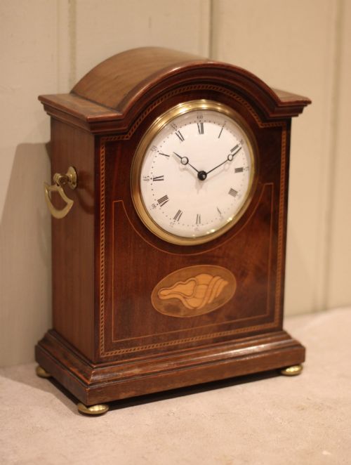 fig 1. An Edwardian arched mahogany mantel clock featuring a shell motif inlay, a white dial with Roman numerals, brass side handles, and brass ball feet.