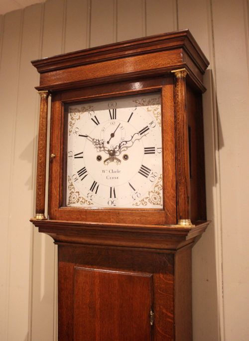 fig 1. A tall late Georgian oak longcase clock featuring a square painted dial, turned hood columns, and a polished wood trunk.
